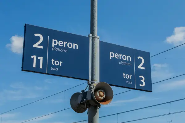 Niebieska tablica informacyjna na dworcu kolejowym wskazuje peron 2, tor 11 oraz peron 2, tor 3. W tle błękitne niebo z chmurami i linie trakcyjne.